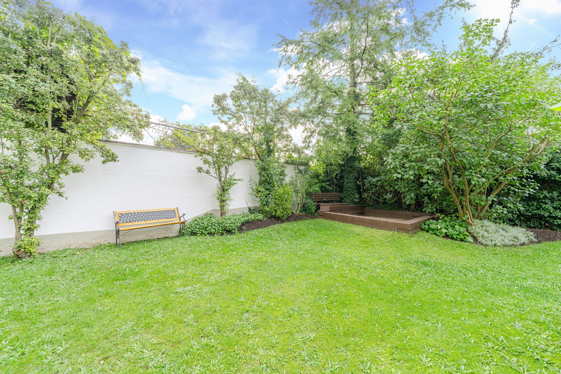 Grüner Garten mit Rasen, Bäumen, Holzbank und Terrasse an einer weißen Mauer.