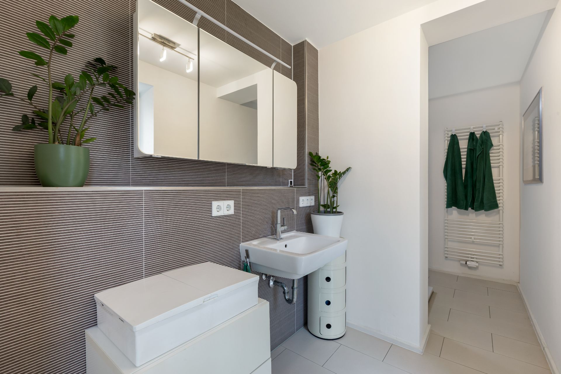 Modernes Badezimmer mit Spiegel, Waschbecken, Pflanzen und Handtuchheizkörper mit grünen Handtüchern.