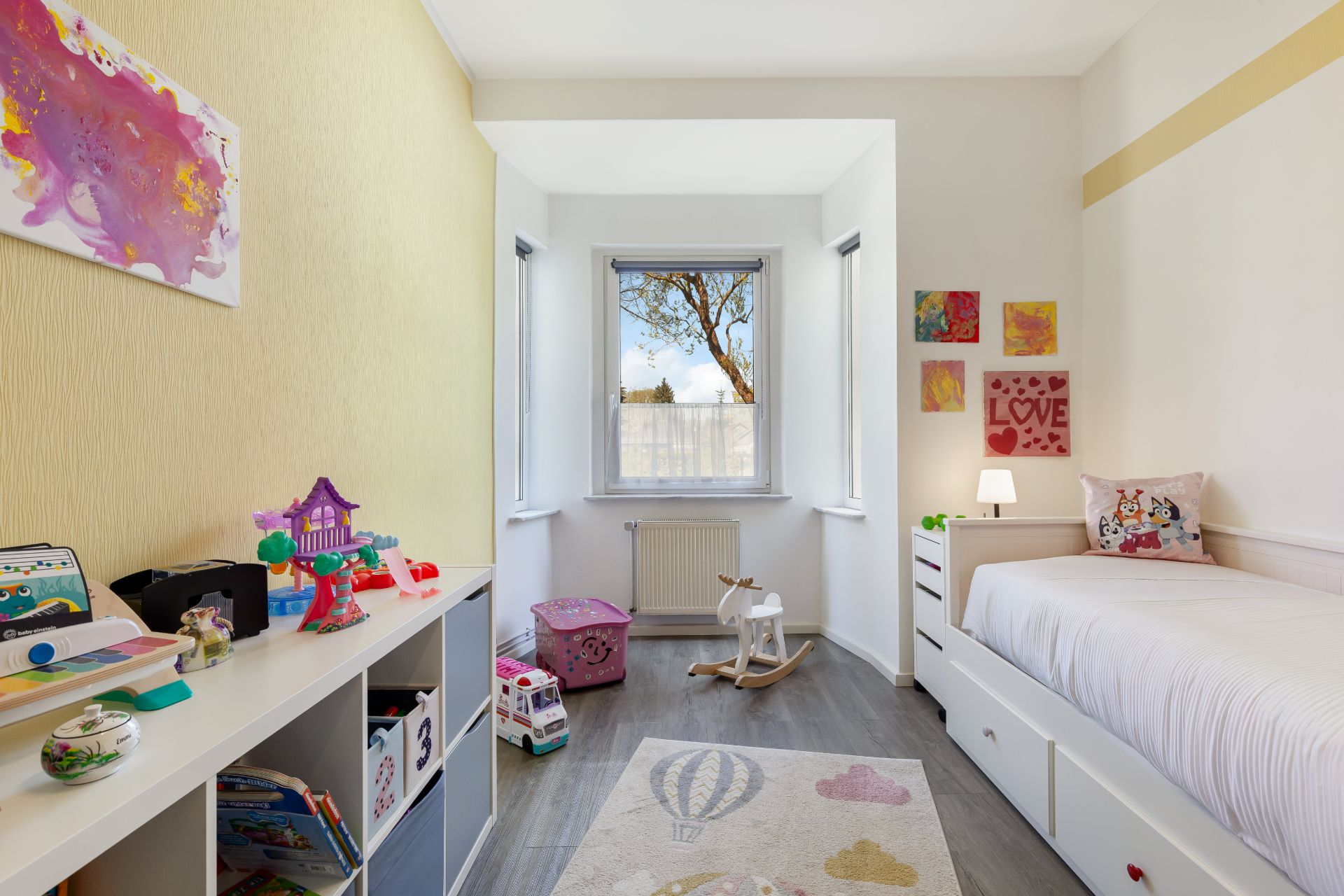 Helles Kinderzimmer mit Einzelbett, Spielsachen, Regal und drei Fenstern.