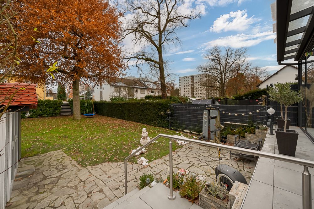 Garten mit Rasen, Bäumen, Spielfläche, Terrasse und Steinweg hinter einem Wohnhaus.