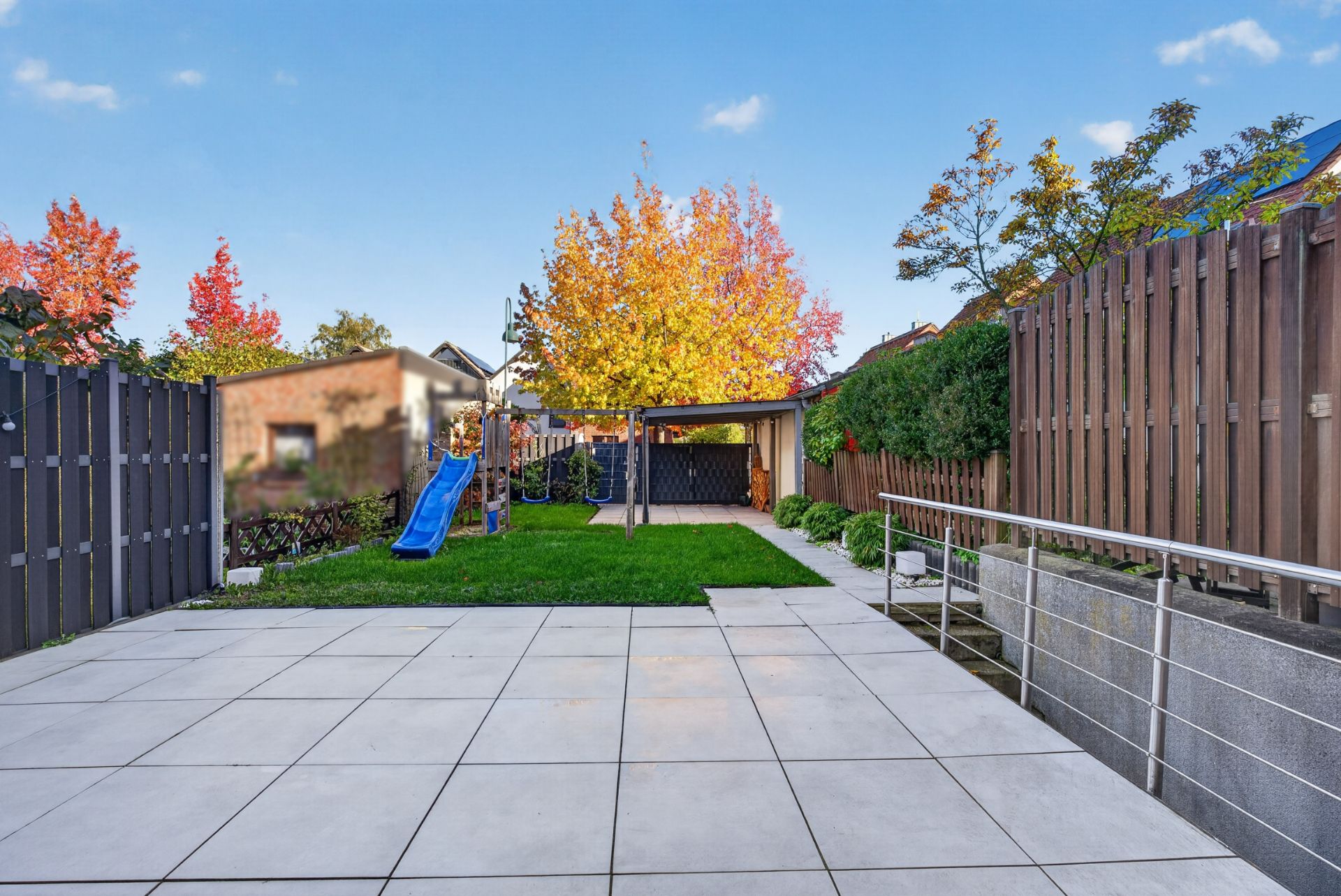 Gepflasterte Terrasse, Rasenfläche, blauer Spielplatz, Holz- und Metallzaun, Bäume im Garten.