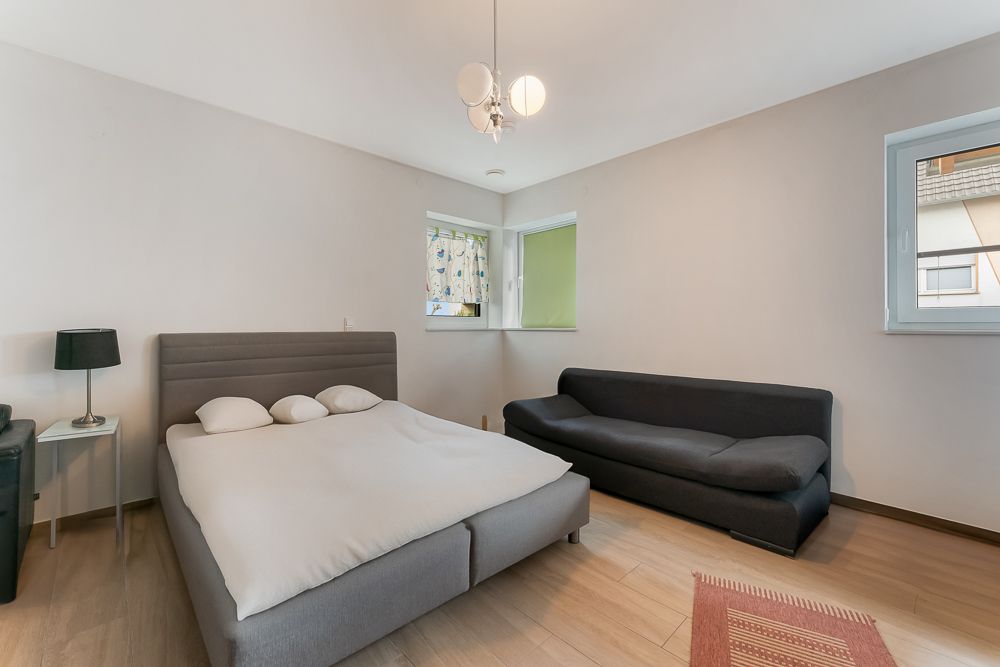 Schlafzimmer mit Doppelbett, schwarzem Sofa, Nachttisch und Lampe, Holzboden und zwei Fenstern.