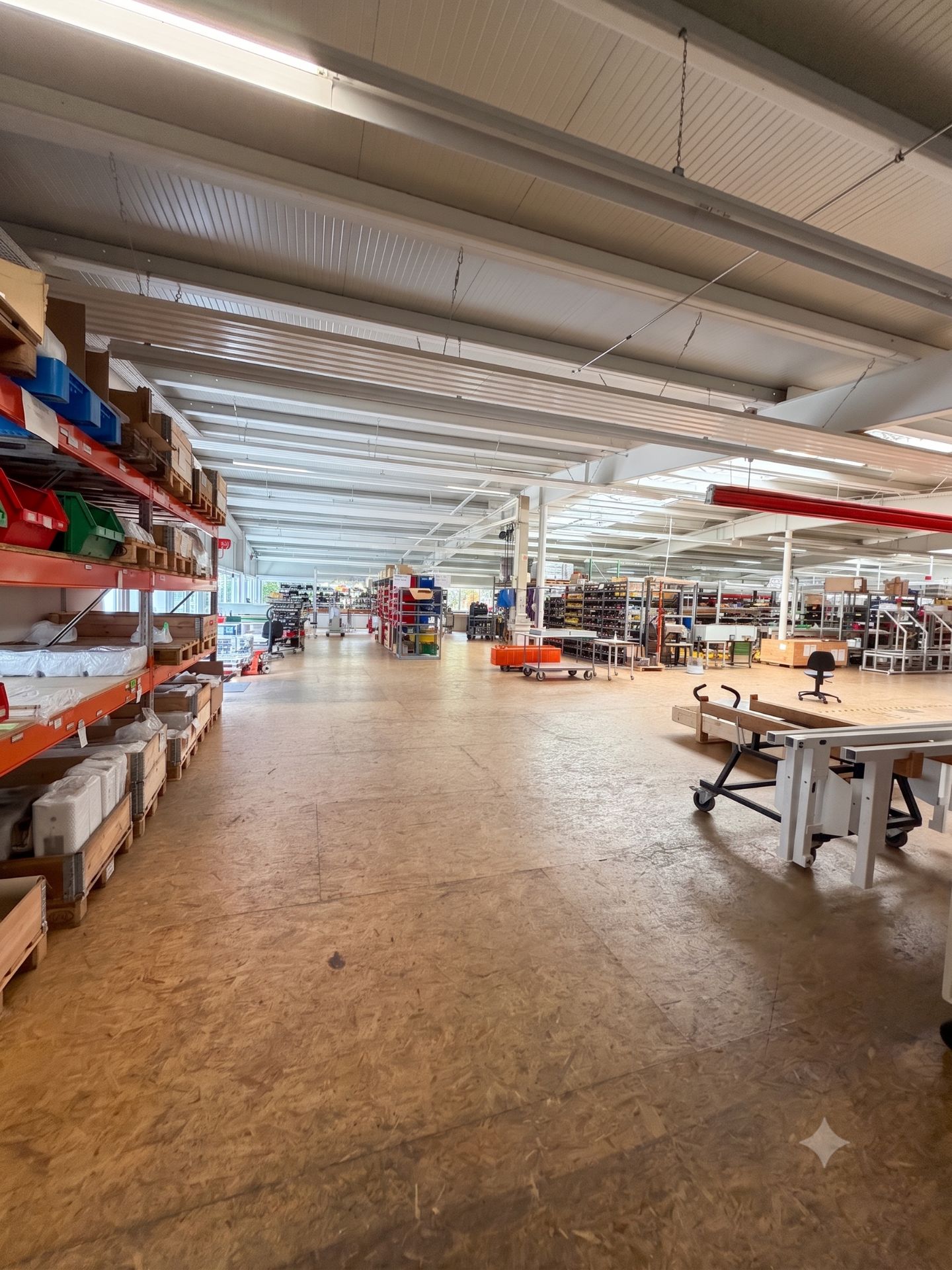 Große Halle mit Regalen, Kartons und Werkbänken, heller Boden und Decke, viele Arbeitsbereiche.