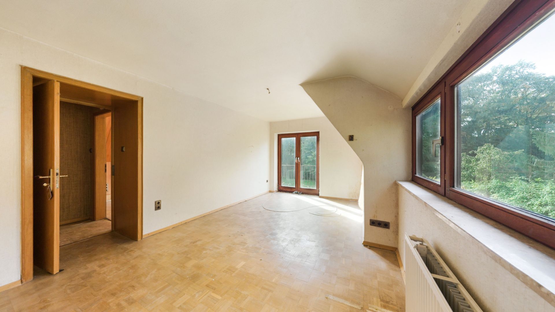 Helles leeres Zimmer mit Parkettboden, großen Fenstern und offen stehender Tür.