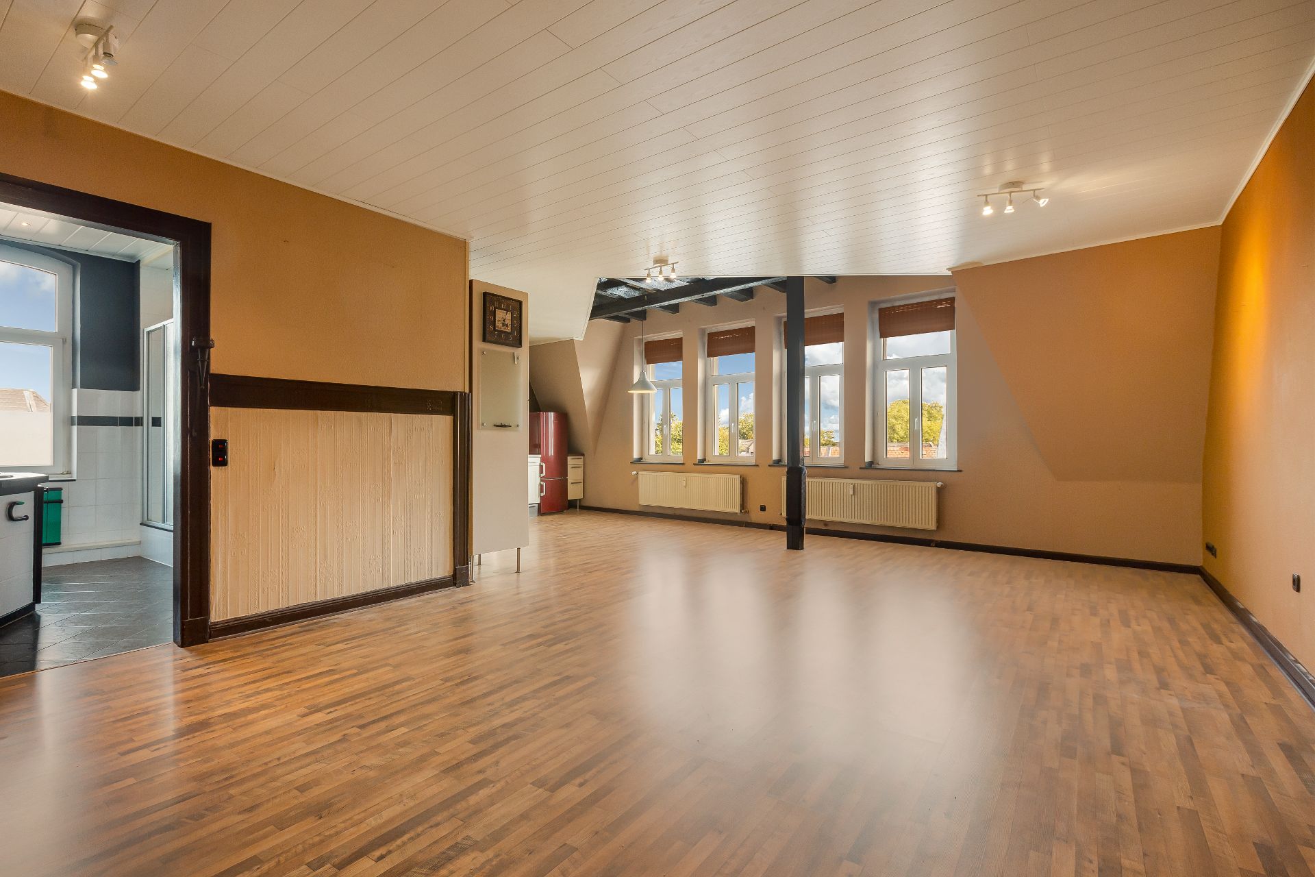 Großes, leeres Wohnzimmer mit Holzfußboden, mehreren Fenstern und angrenzender Küche.