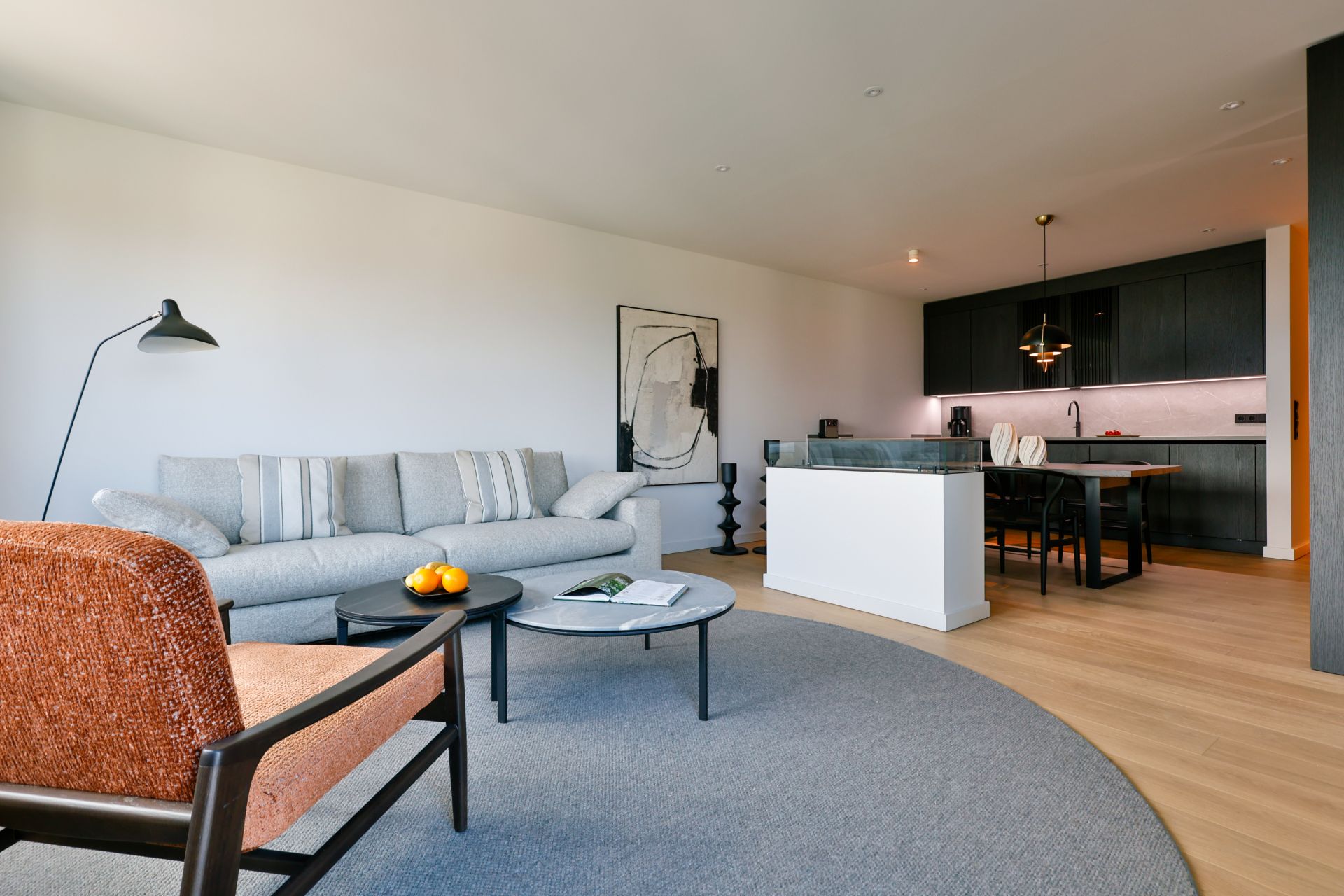 Helles Wohnzimmer mit Sofa, Sessel, Couchtisch und offener moderner Küche im Hintergrund.