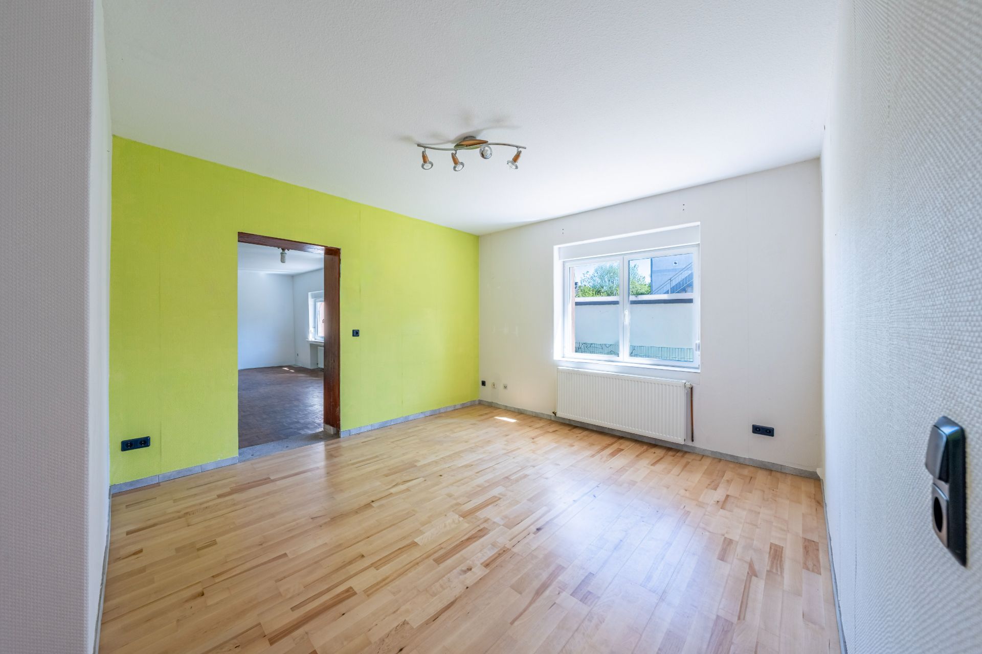 Helles Zimmer mit Holzfußboden, gelber Wand und großem Fenster mit Tageslicht.