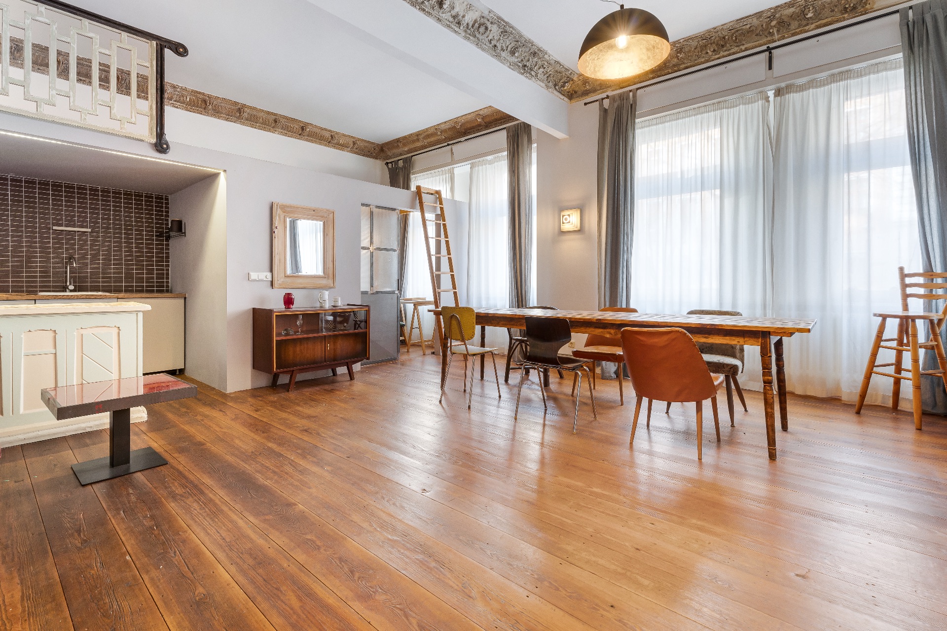 Helles Wohnzimmer mit Holzboden, Esstisch, bunten Stühlen, Küchenzeile und großen Fenstern mit Vorhängen.