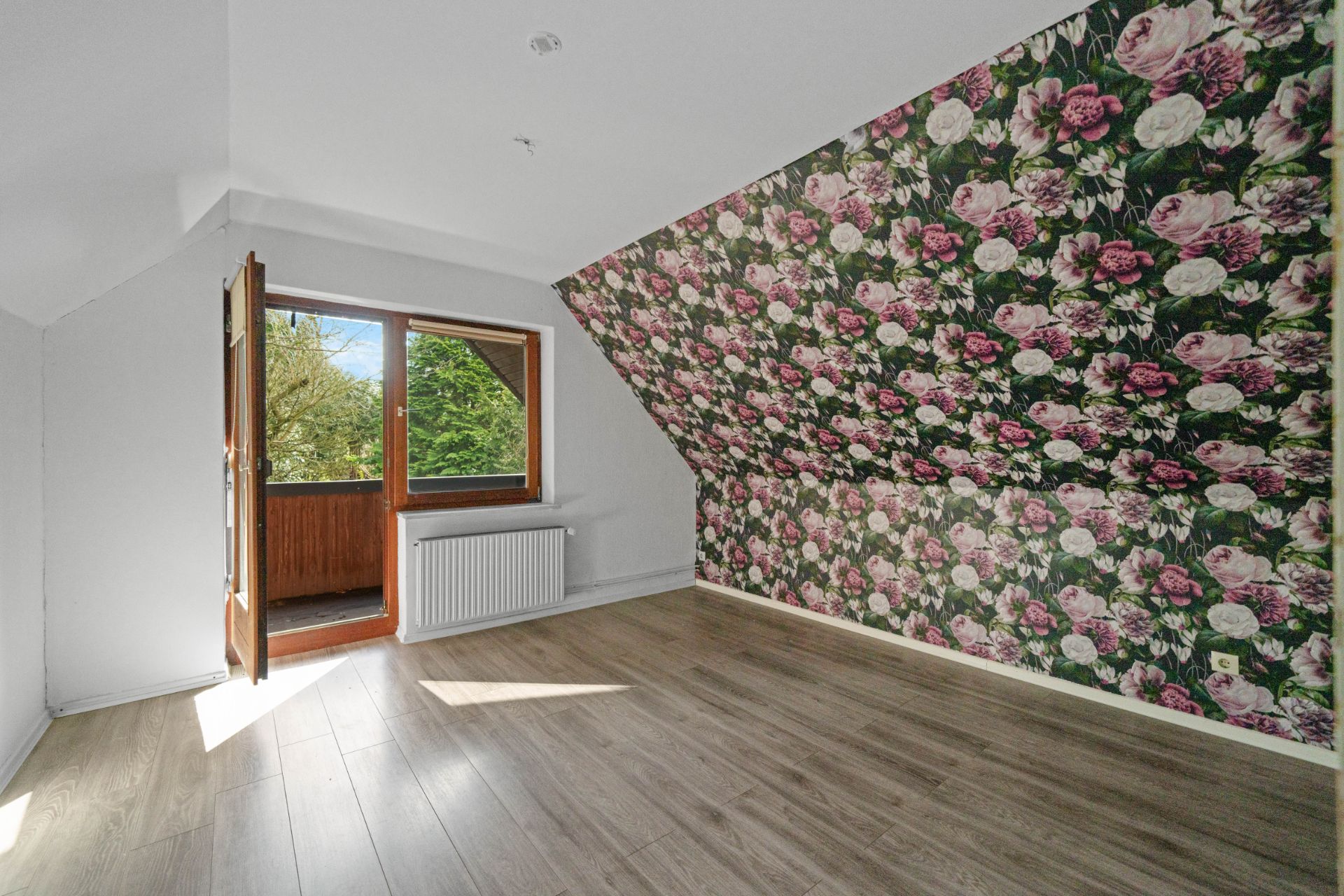 Leeres Zimmer mit schräger Wand, bunter Blumen-Tapete und offenem Fenster zum Balkon.