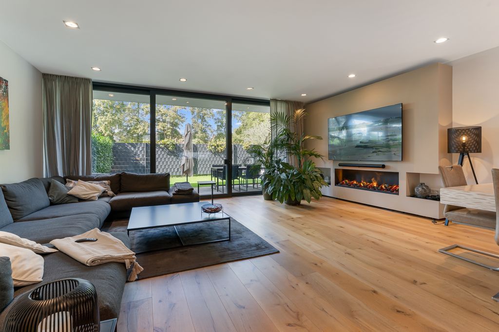 Modernes Wohnzimmer mit großem Sofa, Kamin, Fernseher und Blick auf Garten durch Glasfront.