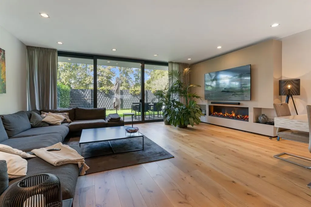Modernes Wohnzimmer mit großem Sofa, Kamin, Fernseher und Blick auf Garten durch Glasfront.