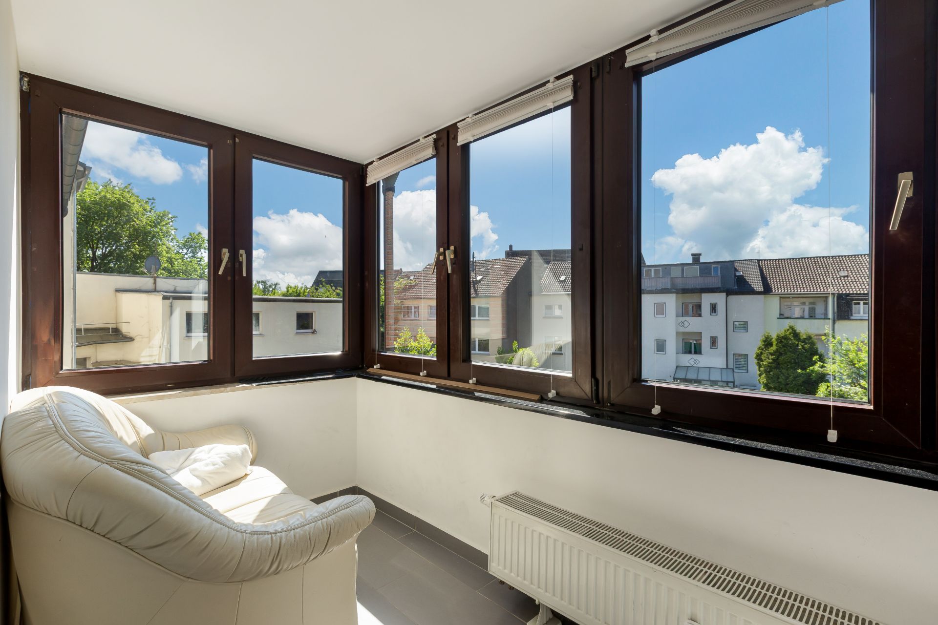 Heller Raum mit großen Fenstern, Sessel und Heizkörper, Blick auf Nachbarhäuser.