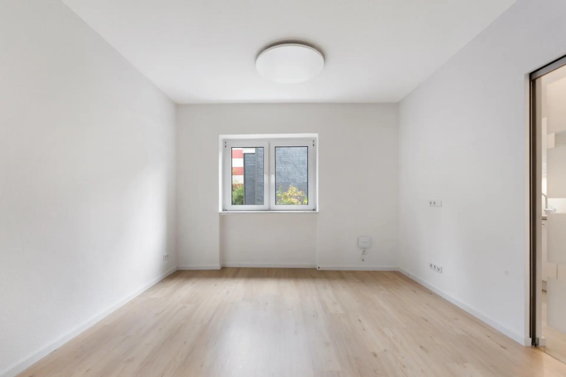 Heller, leerer Raum mit Holzfußboden, weißem Fenster und Deckenleuchte.