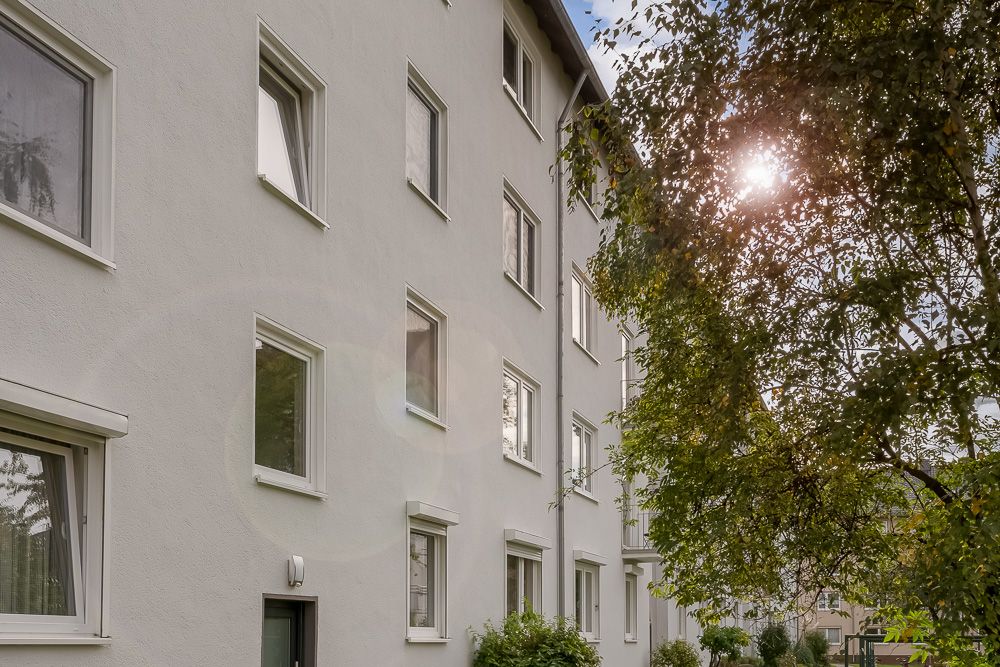 Mehrfamilienhaus mit mehreren Fenstern und Bäumen im Vordergrund bei Sonnenschein.