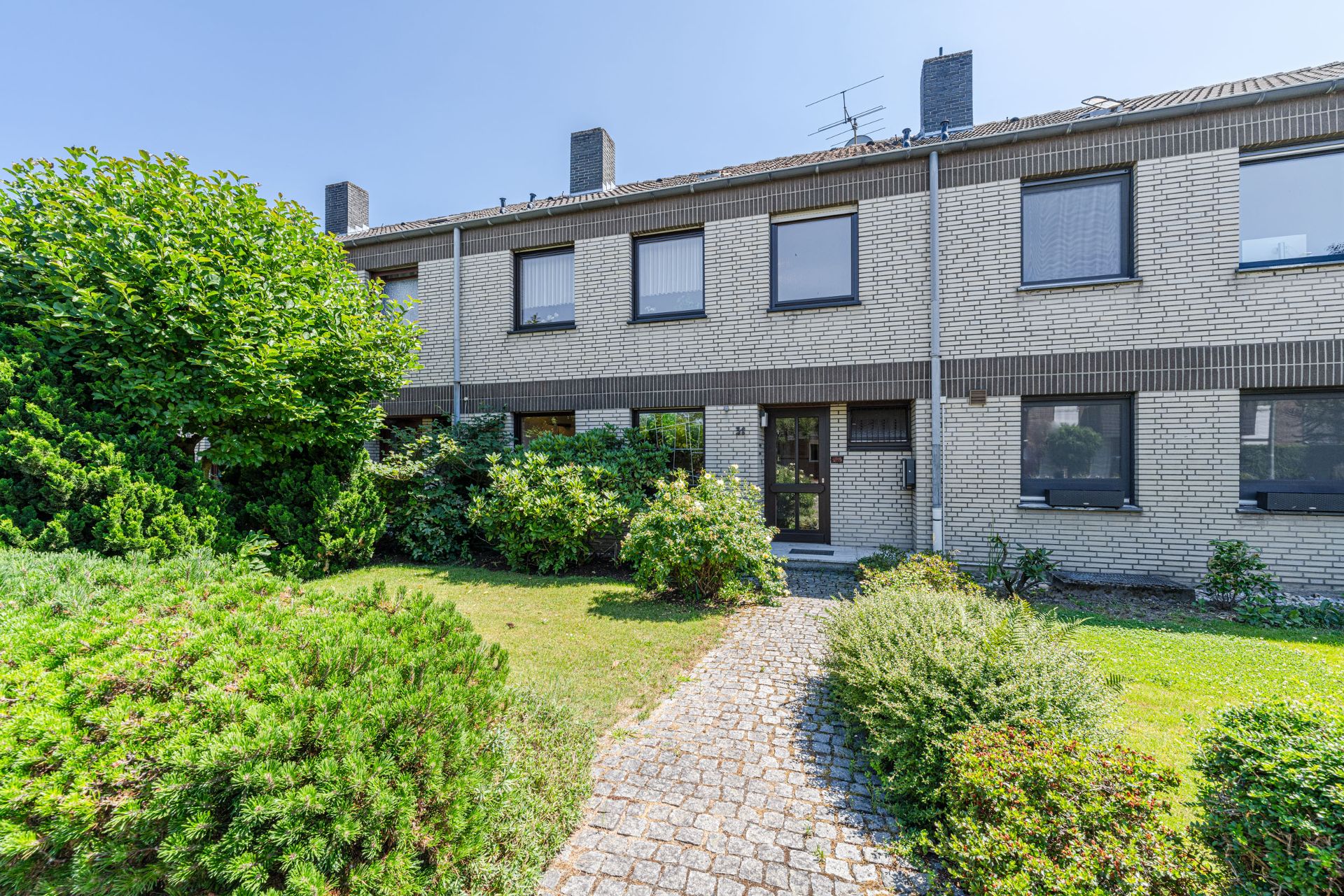 Zweistöckiges Reihenhaus mit Vorgarten und vielen Büschen an einem sonnigen Tag.