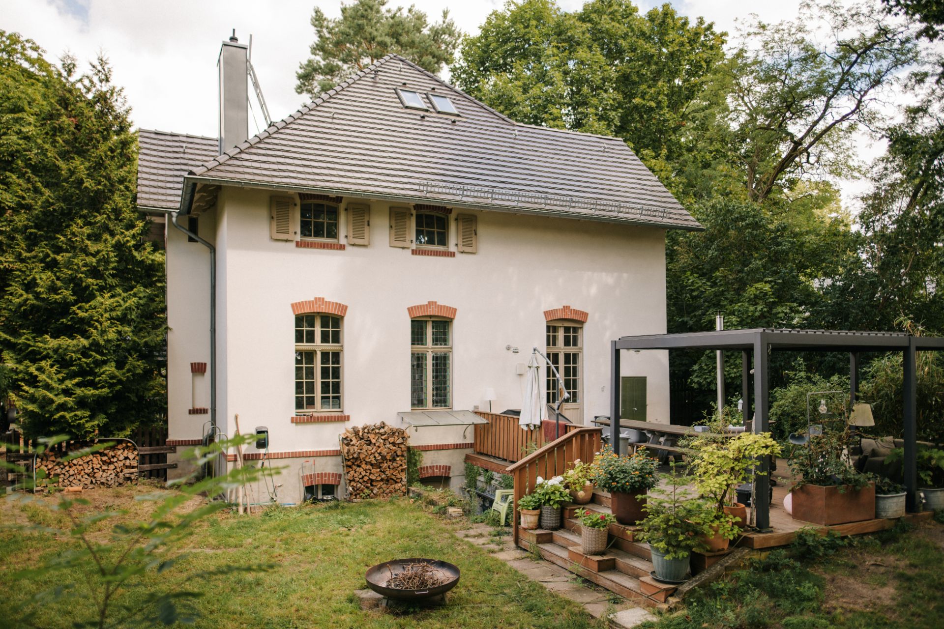 Weißes Haus mit großem Garten, Terrasse, Holzstapeln und vielen Pflanzen, umgeben von Bäumen.