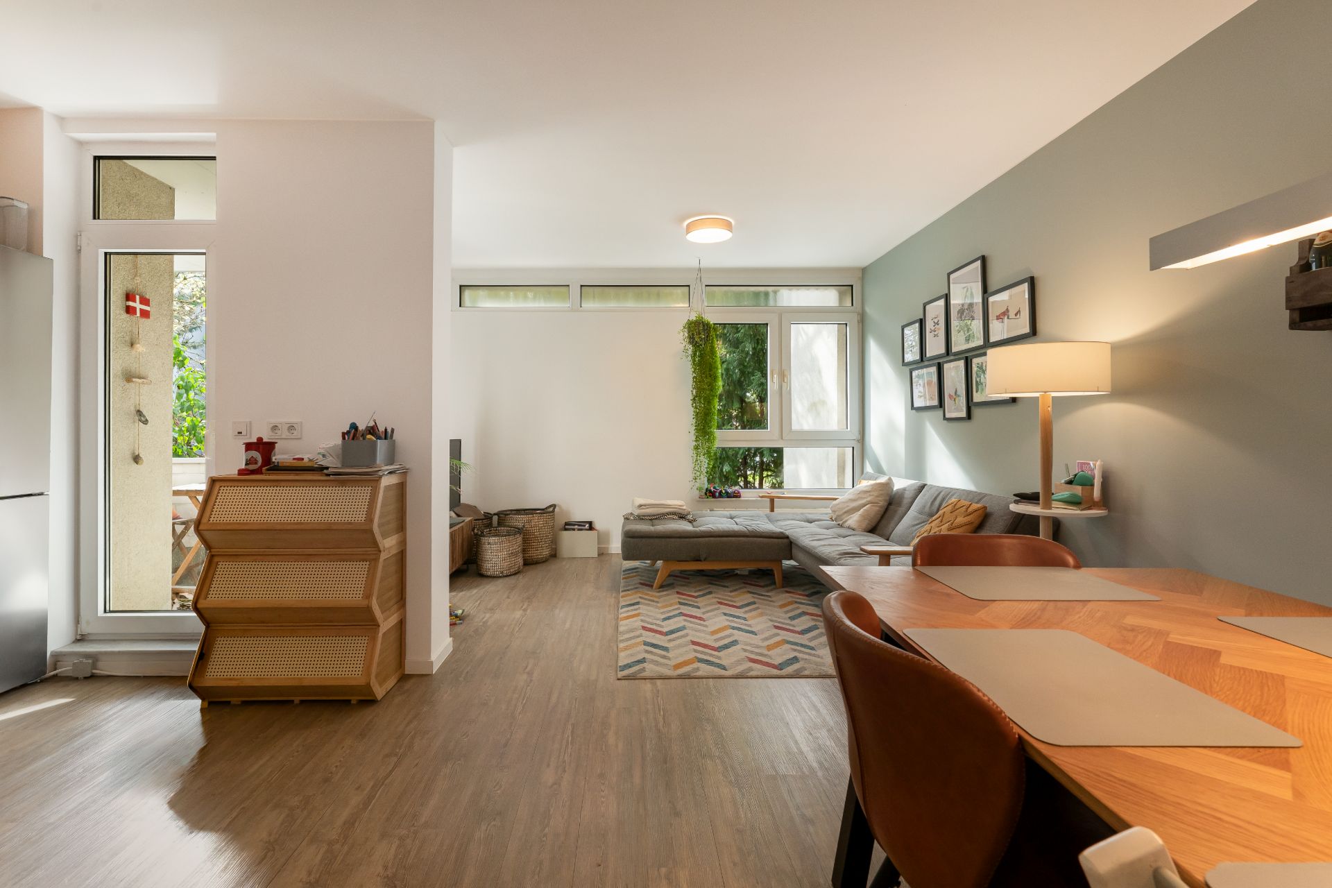Helles Wohnzimmer mit Sofa, Esstisch, Wandbildern und großen Fenstern.