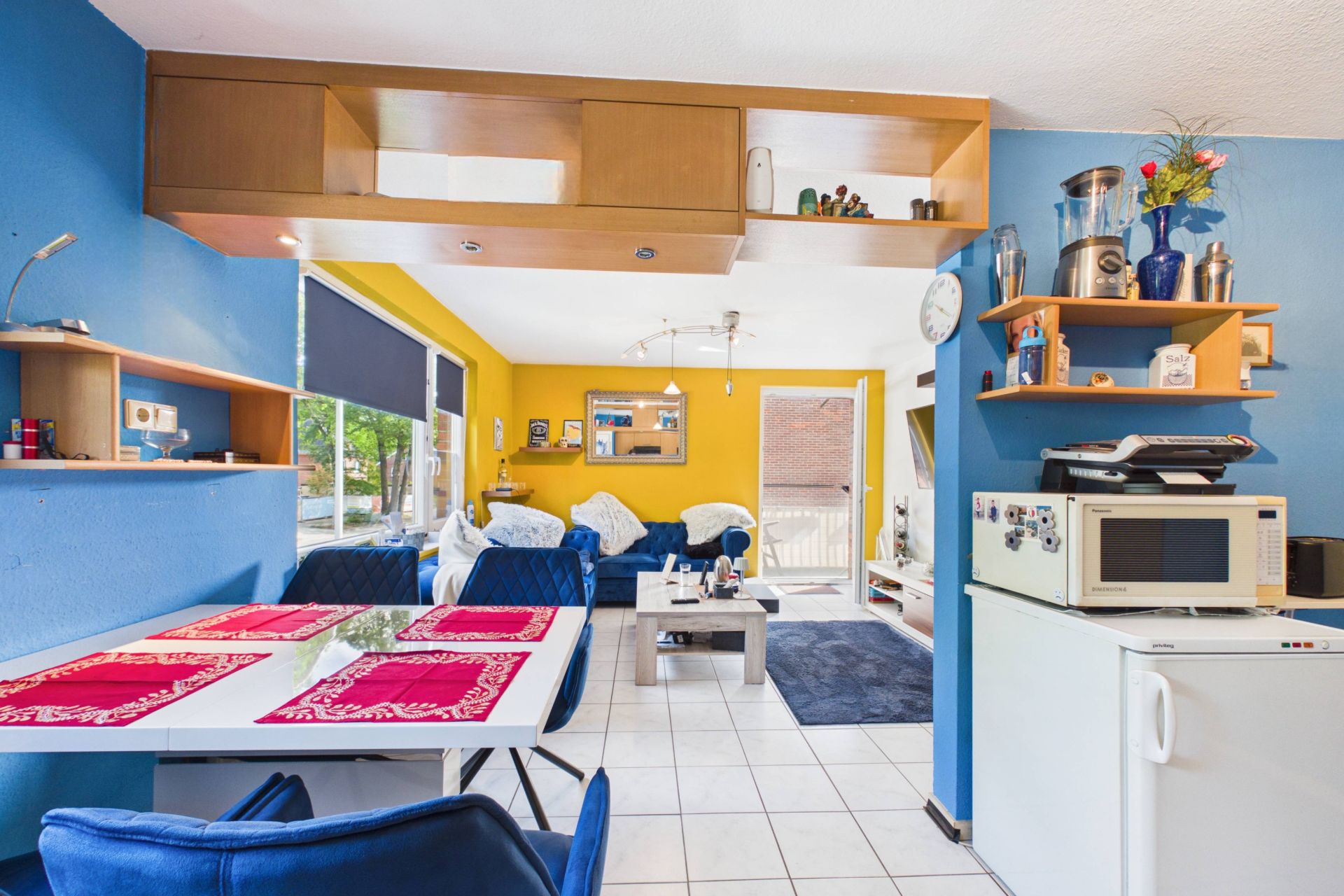 Offener Wohnraum mit Essecke, Sofa, Couchtisch, Kühlschrank und Mikrowelle, gelben und blauen Wänden.