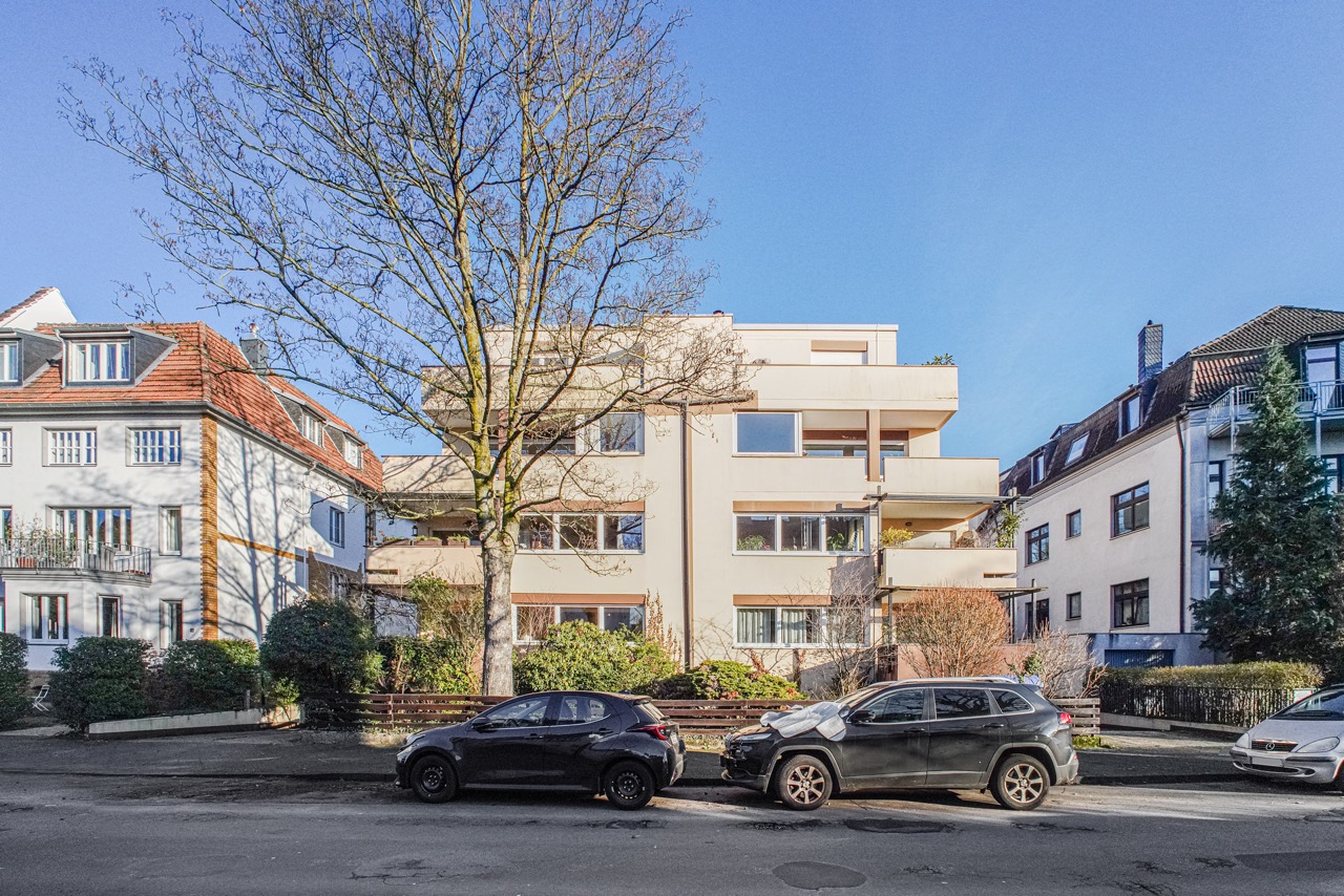 Wohngebäude mit Balkonen, davor parken Autos, ein großer Baum steht an der Straße.