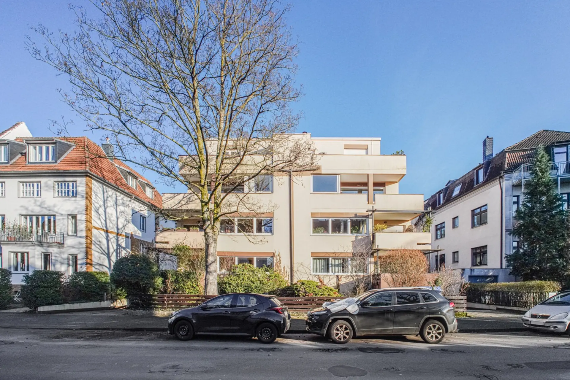 Wohngebäude mit Balkonen, davor parken Autos, ein großer Baum steht an der Straße.