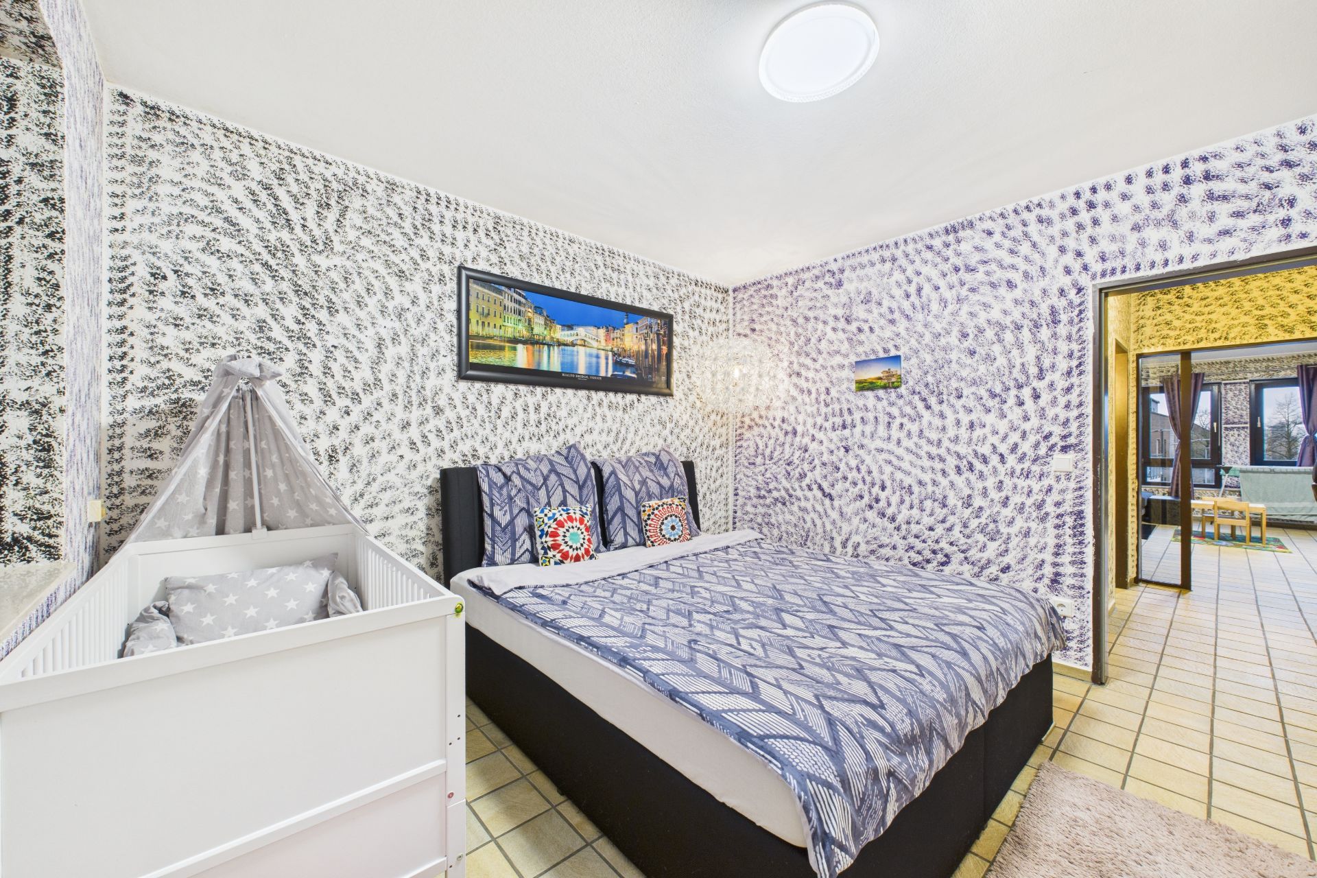Schlafzimmer mit Doppelbett, Babybett und gemusterten Wänden.