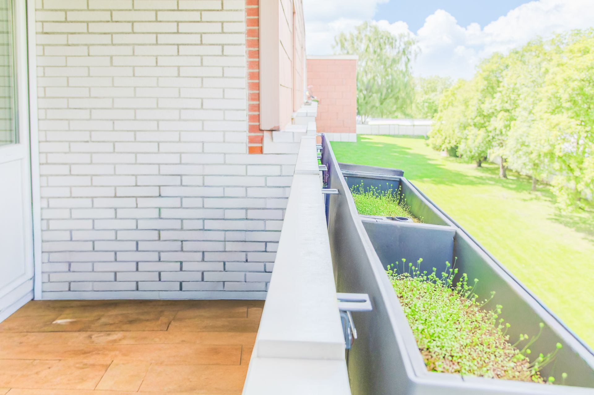 Balkon mit weißen Ziegelwänden, Holzboden und Pflanzkästen, Blick auf grünen Garten.