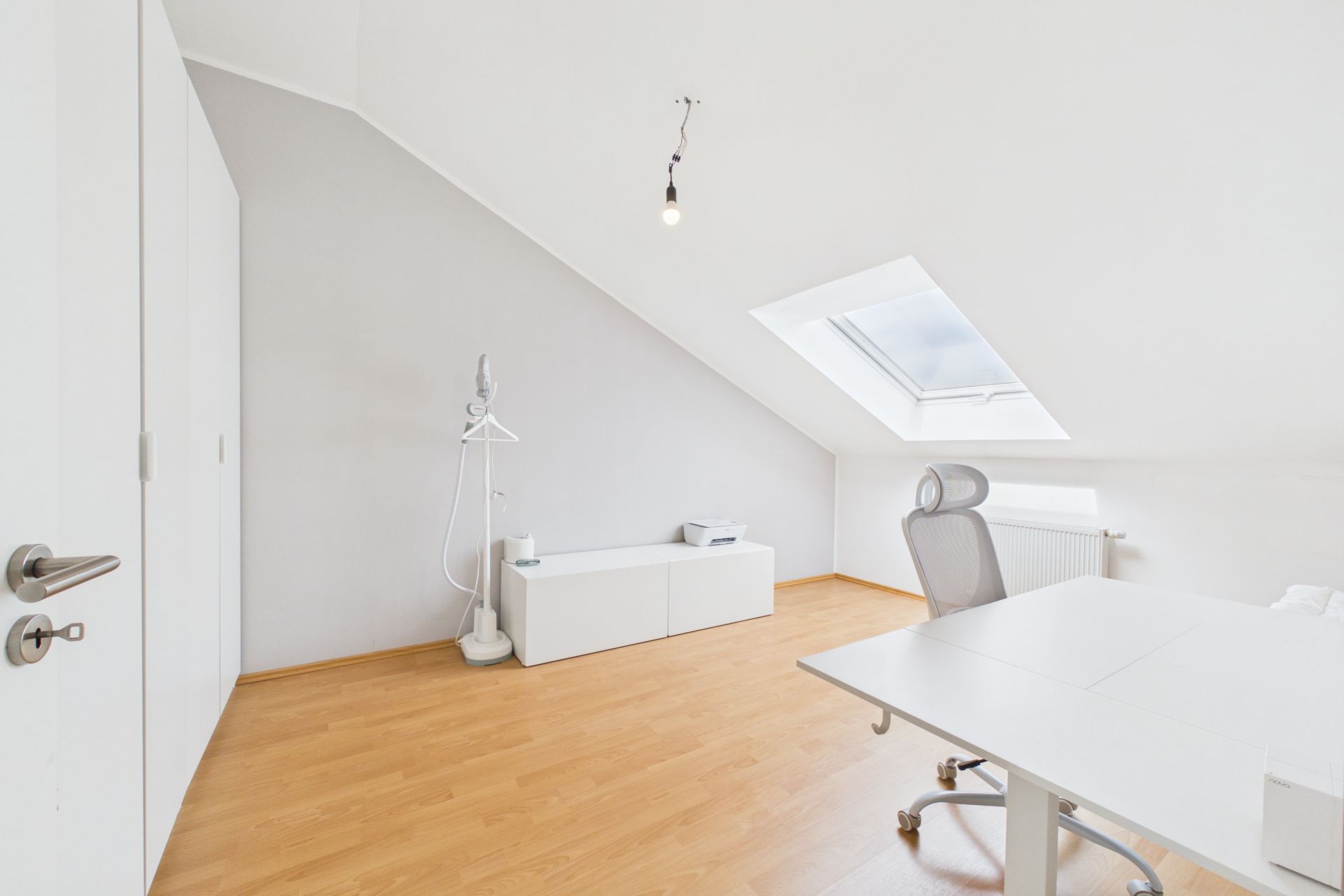 Helles Zimmer mit Schreibtisch, Bürostuhl, Sideboard und Dachfenster.