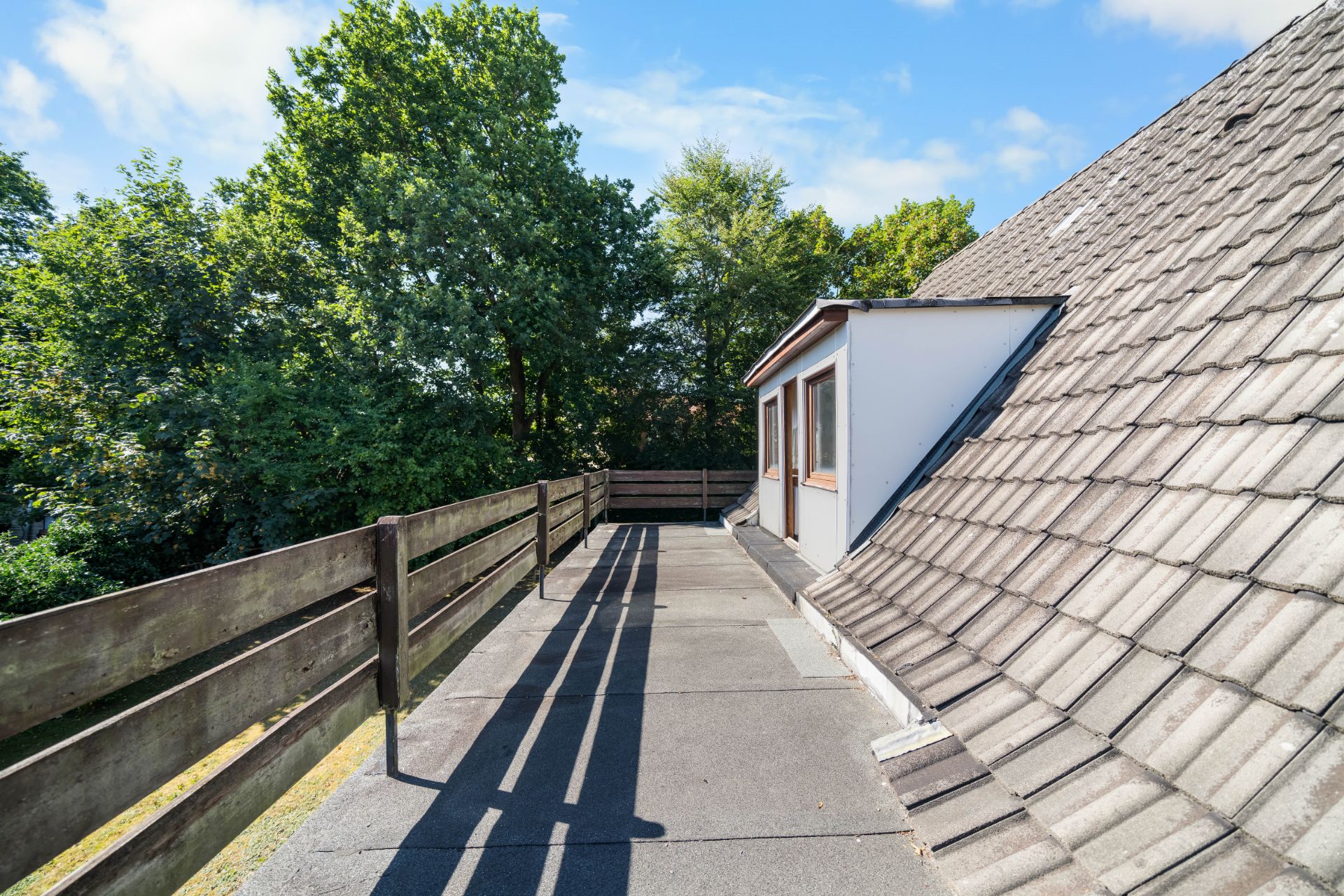Große Dachterrasse mit Holzzaun und Blick auf Bäume neben einem Haus mit Schrägdach.
