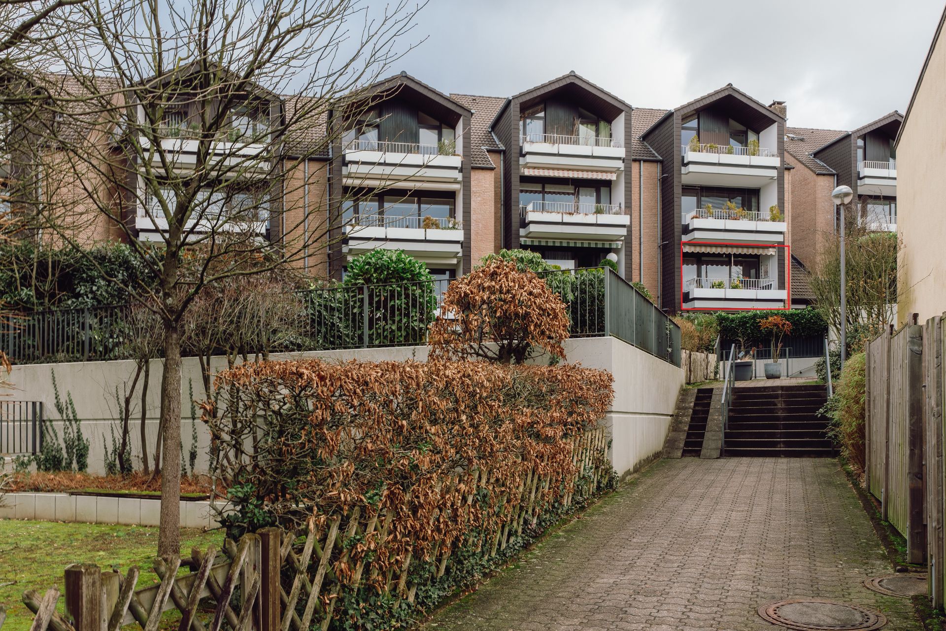 Mehrfamilienhaus mit Balkonen, Hecken und kahlen Bäumen im Vordergrund.