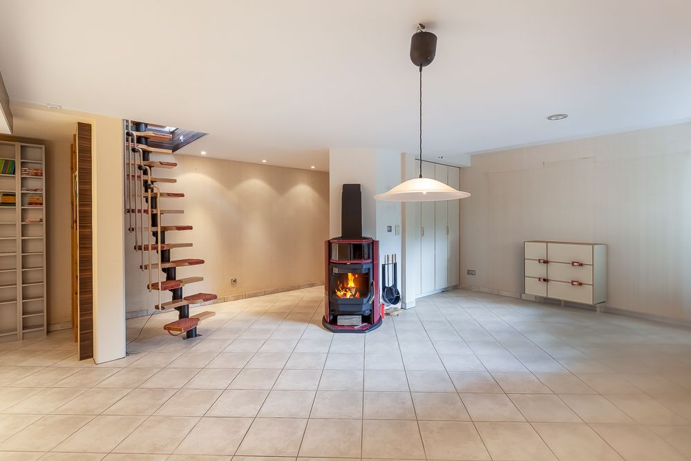 Helles Wohnzimmer mit Fliesenboden, Holztreppe, Kaminofen und Schränken.