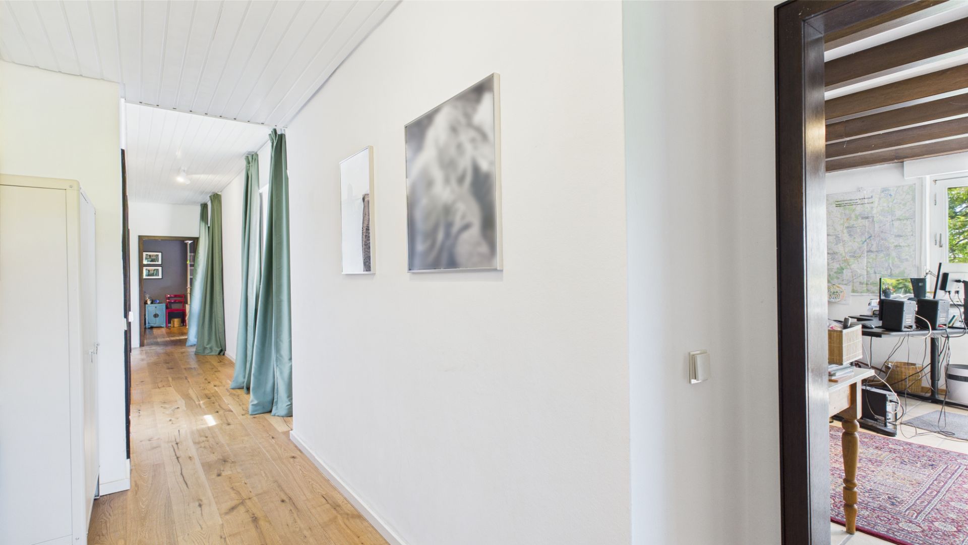 Heller Flur mit Holzboden, weißen Wänden, zwei Bildern und Blick in ein Arbeitszimmer.