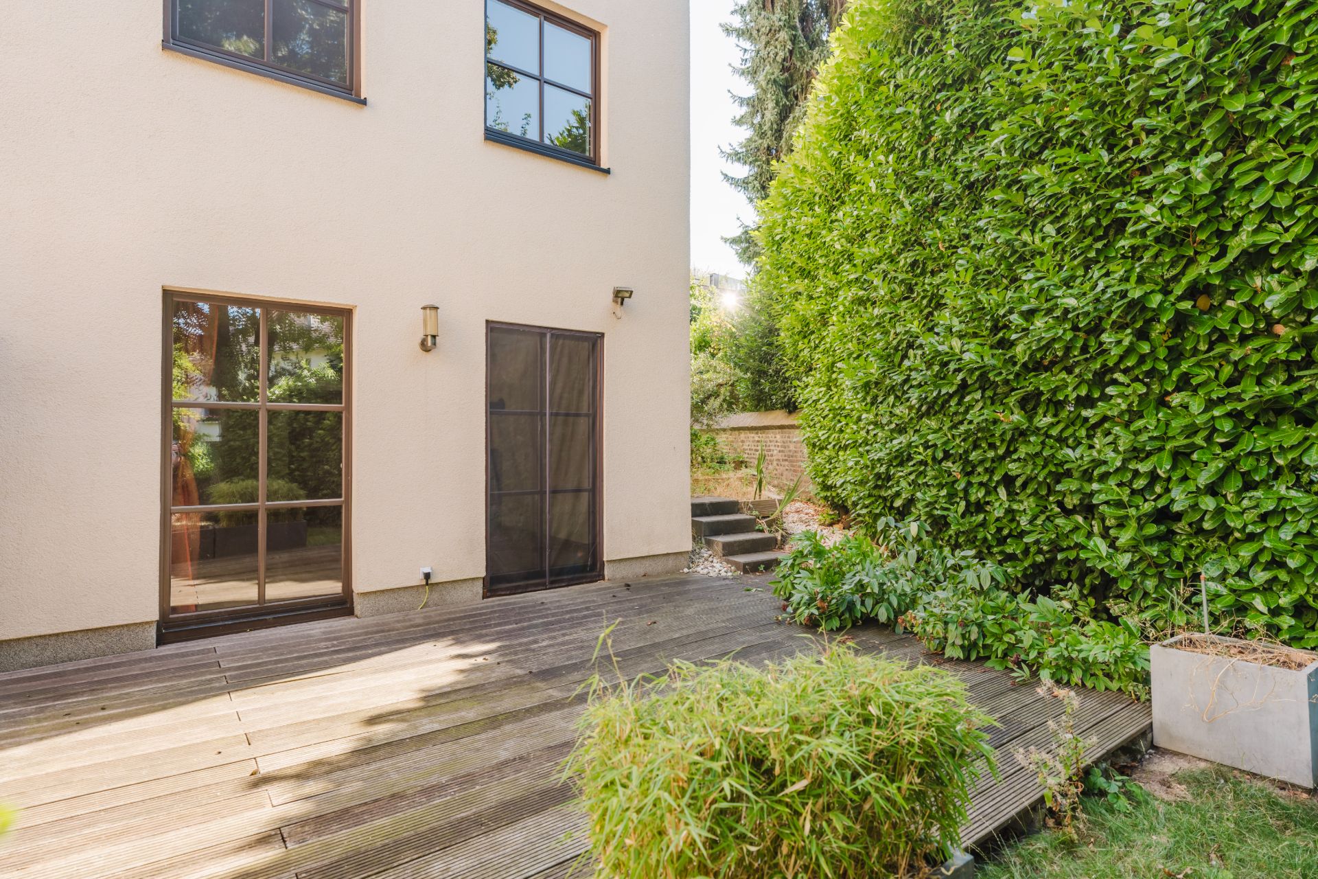Holzterrasse mit Pflanzen neben einem modernen Wohnhaus und einer hohen grünen Hecke.