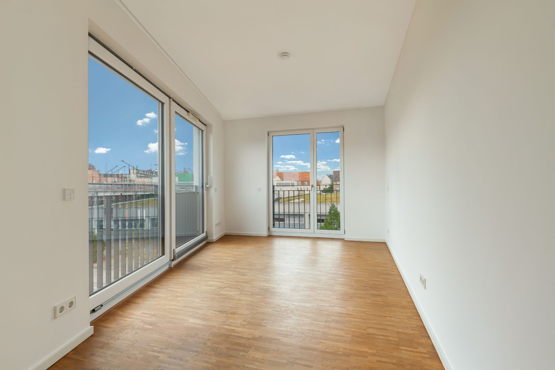Helles, leeres Zimmer mit Holzboden und großen Fenstern, die auf einen Balkon führen.