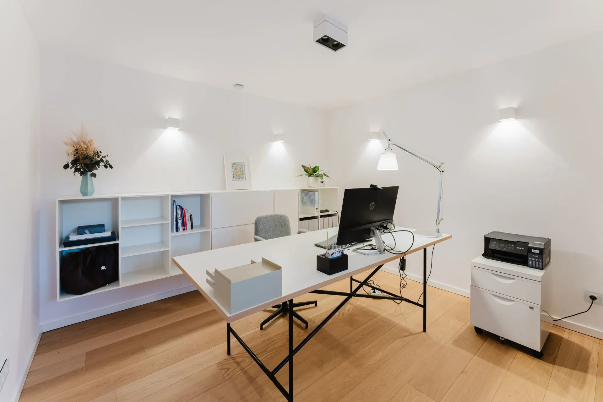 Modernes Arbeitszimmer mit Schreibtisch, Computer, Regal, Lampe und Drucker auf Holzfußboden.