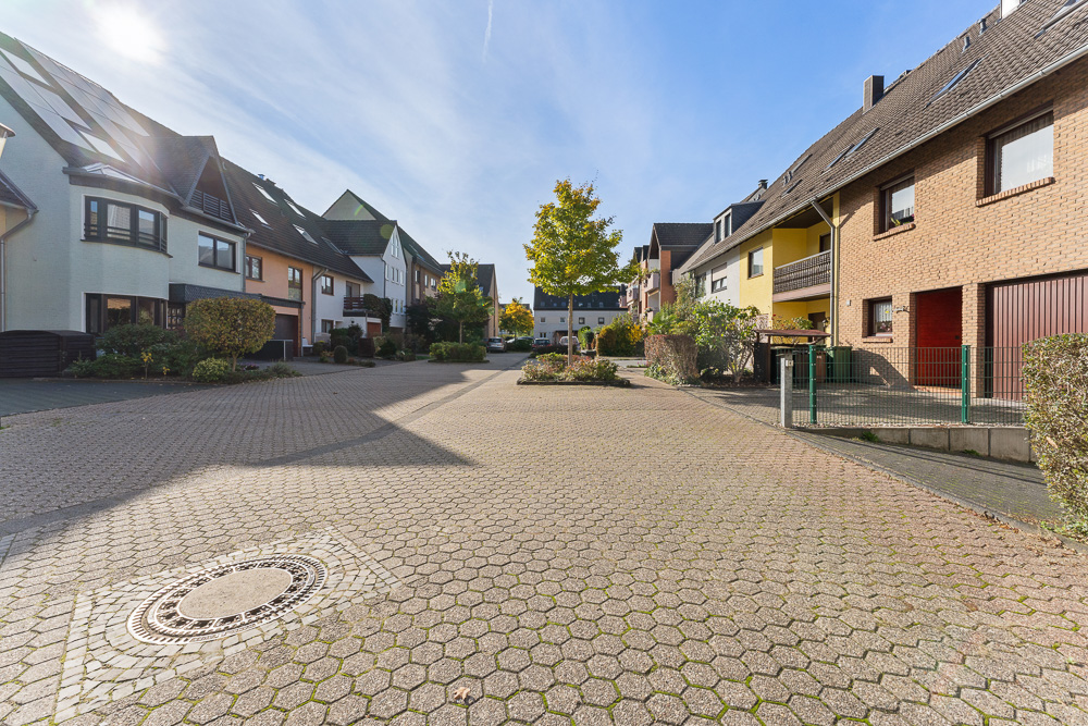 Gepflasterte Wohnstraße mit Reihenhäusern und Bäumen bei sonnigem Wetter.
