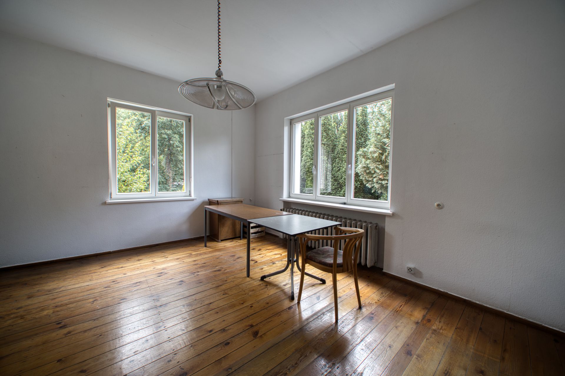 Helles Zimmer mit Holzboden, zwei Fenstern, zwei Tischen und zwei Stühlen.