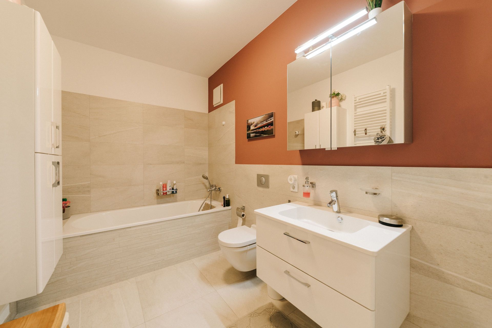 Modernes Badezimmer mit Badewanne, Waschbecken, WC und großem Spiegel an einer rot gestrichenen Wand.