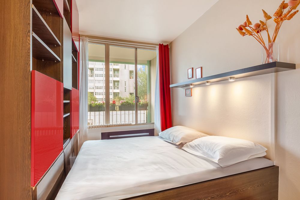 Helles Schlafzimmer mit Doppelbett, Regalwand und großem Fenster mit rotem Vorhang.