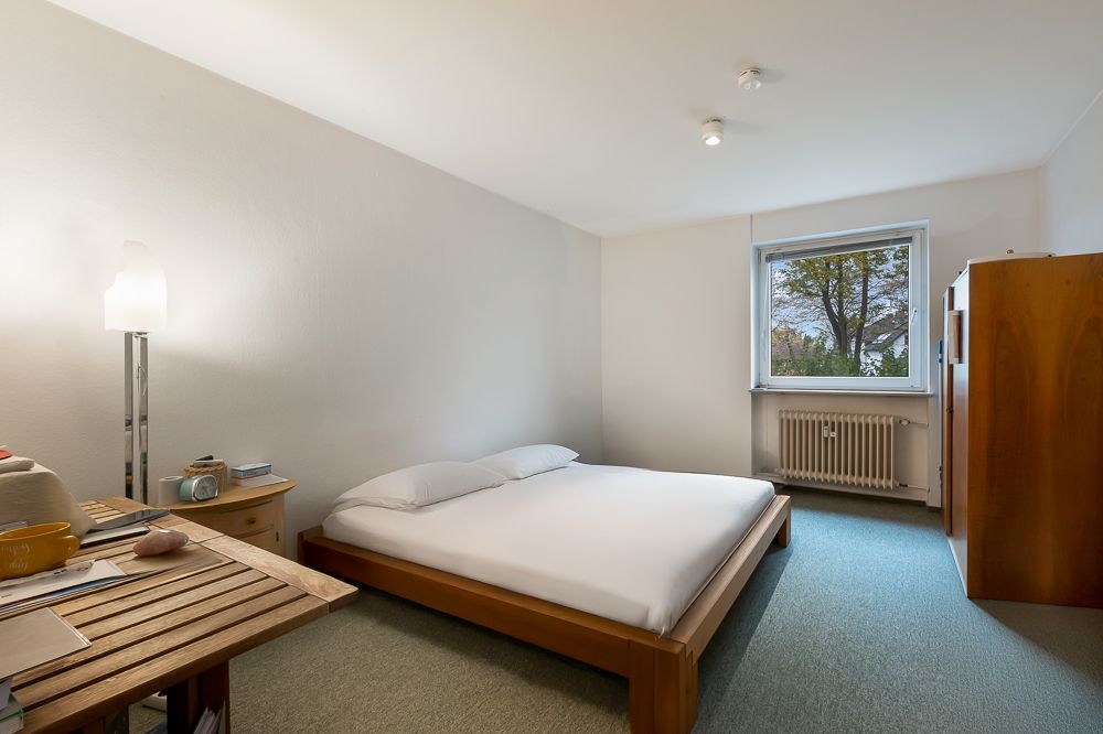 Schlafzimmer mit niedrigem Bett, Schreibtisch, Nachttisch, Kleiderschrank und Fenster mit Blick nach draußen.