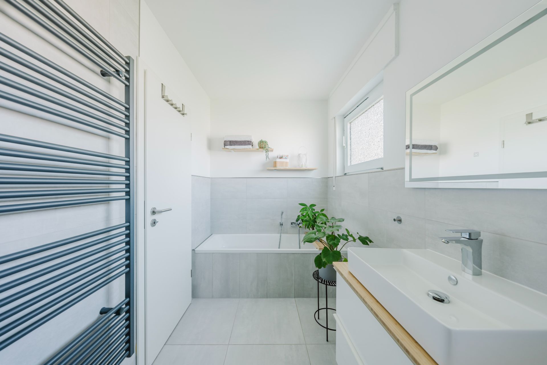 Modernes Badezimmer mit Badewanne, Waschbecken, Spiegel, Handtuchheizkörper und Pflanze.