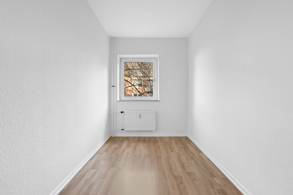 Leerer, schmaler Raum mit weiß gestrichenen Wänden, Holzboden und Fenster mit Heizkörper darunter.