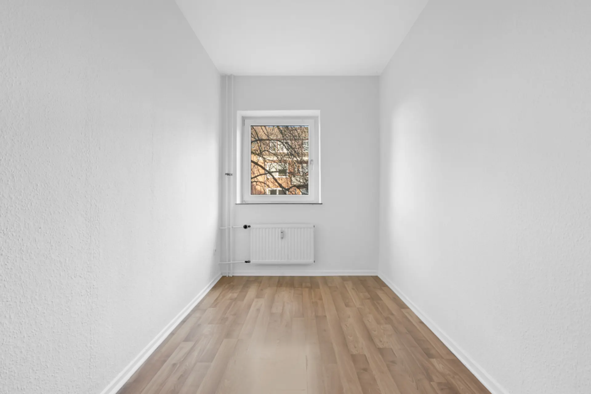Leerer, schmaler Raum mit weiß gestrichenen Wänden, Holzboden und Fenster mit Heizkörper darunter.
