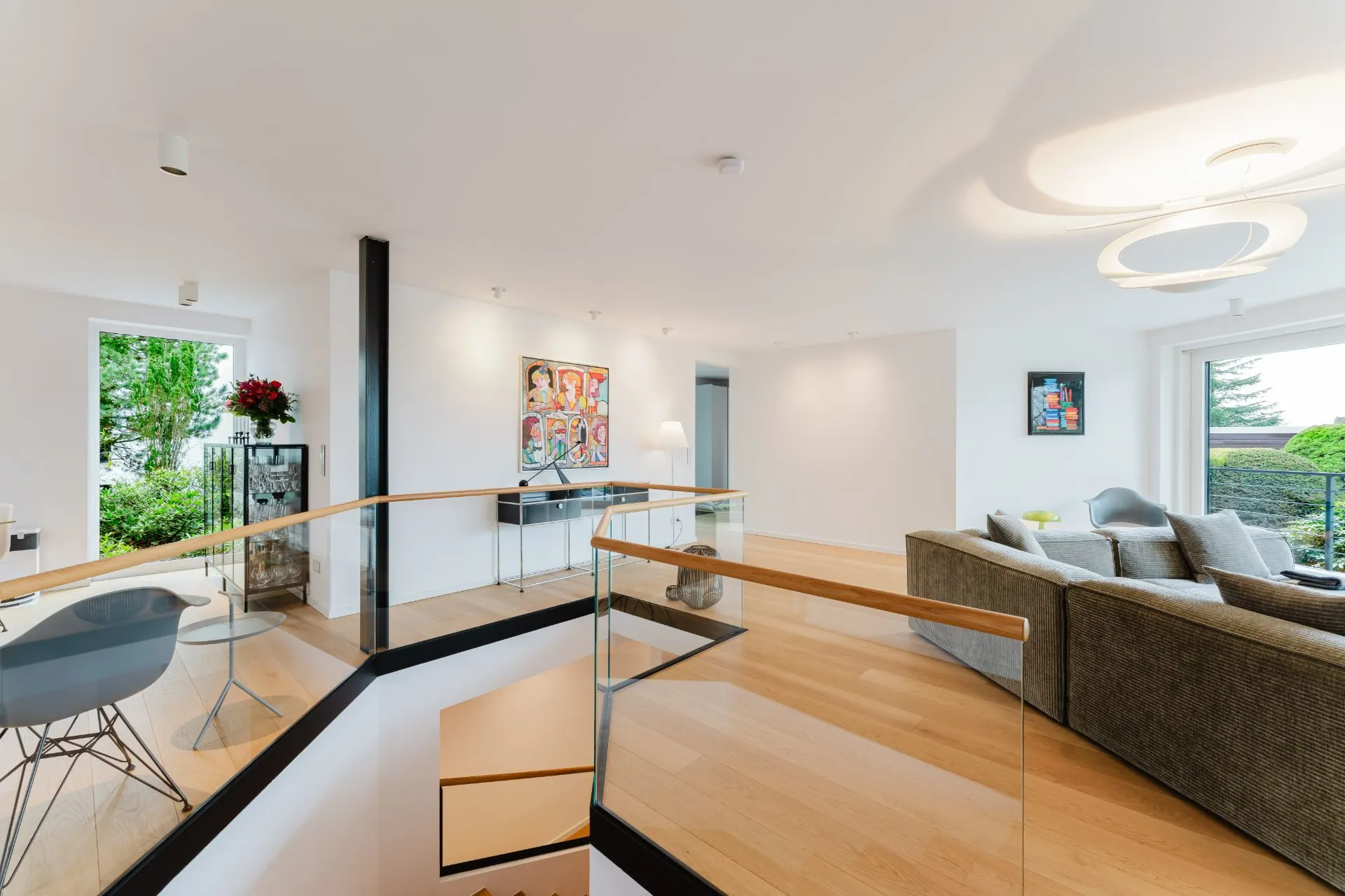 Modernes Wohnzimmer mit grauem Sofa, Glastreppe und großem Fenster mit Blick ins Grüne.