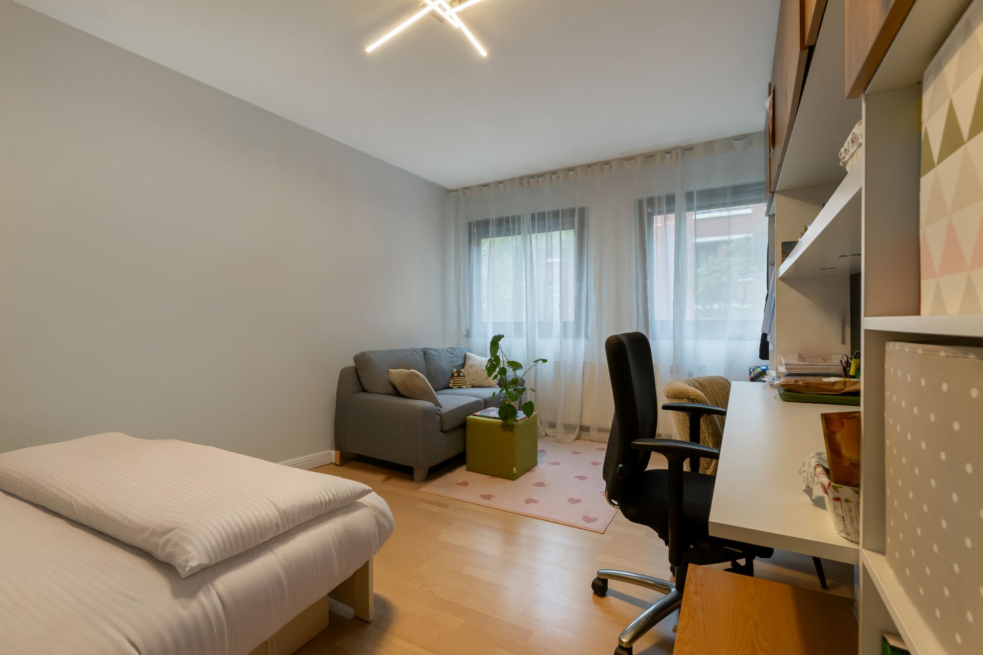 Helles Zimmer mit Bett, Schreibtisch, Bürostuhl, Sofa und zwei Fenstern mit Gardinen.