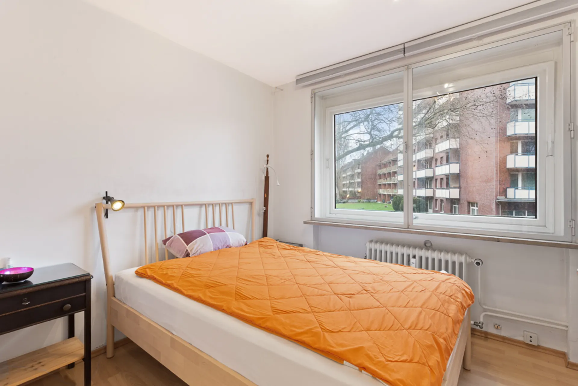 Schlafzimmer mit Einzelbett, Nachttisch und großem Fenster mit Blick auf Wohngebäude.