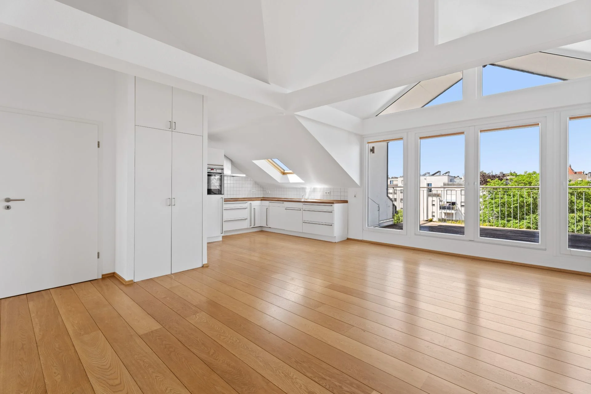 Helles Wohnzimmer mit offener Küche, Parkettboden und großen Fenstern mit Blick auf einen Balkon.