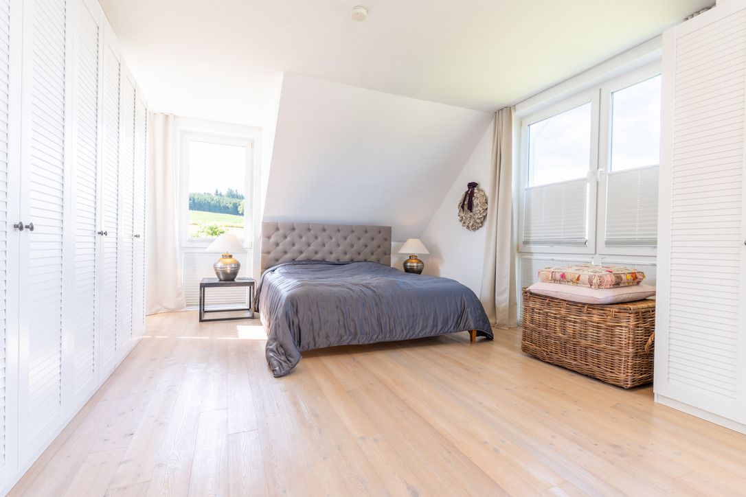 Helles Schlafzimmer mit Doppelbett, großen Fenstern, Einbauschränken und Korbtruhe auf hellem Holzboden.