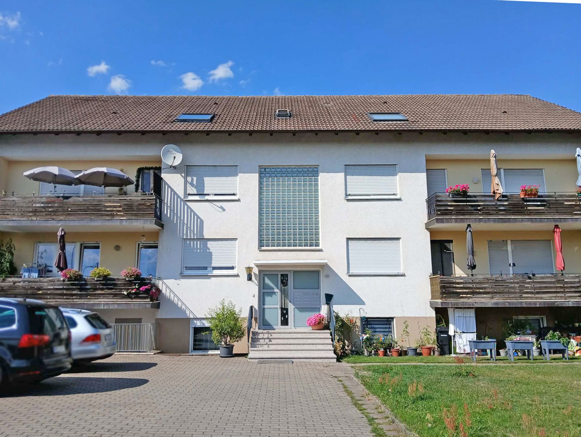 Mehrfamilienhaus mit Balkonen, Pflanzen und parkenden Autos vor dem Eingang.
