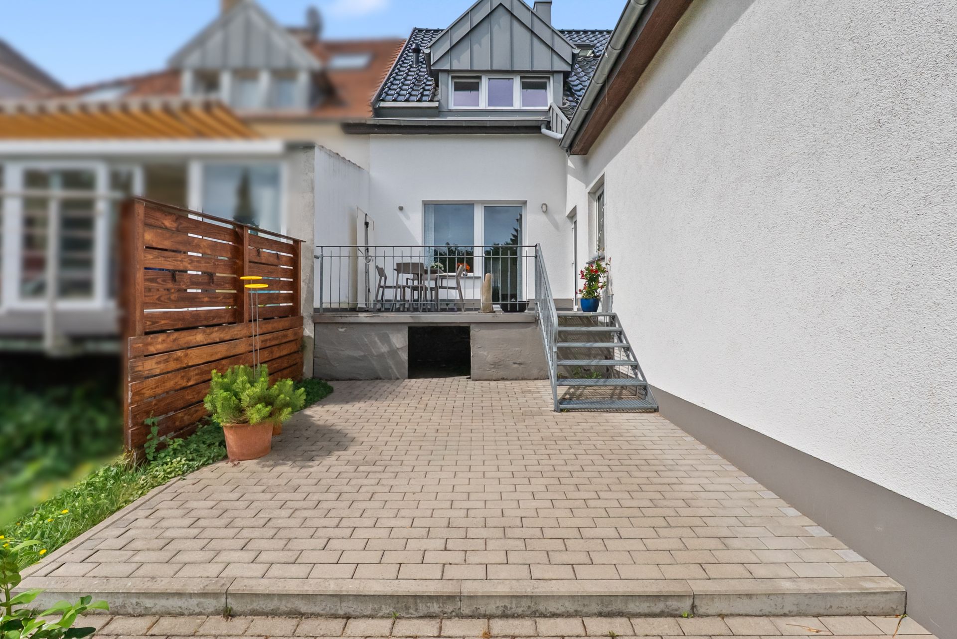 Gepflasterte Terrasse mit Treppe, Geländer und einem Tisch mit Stühlen vor einem Wohnhaus.