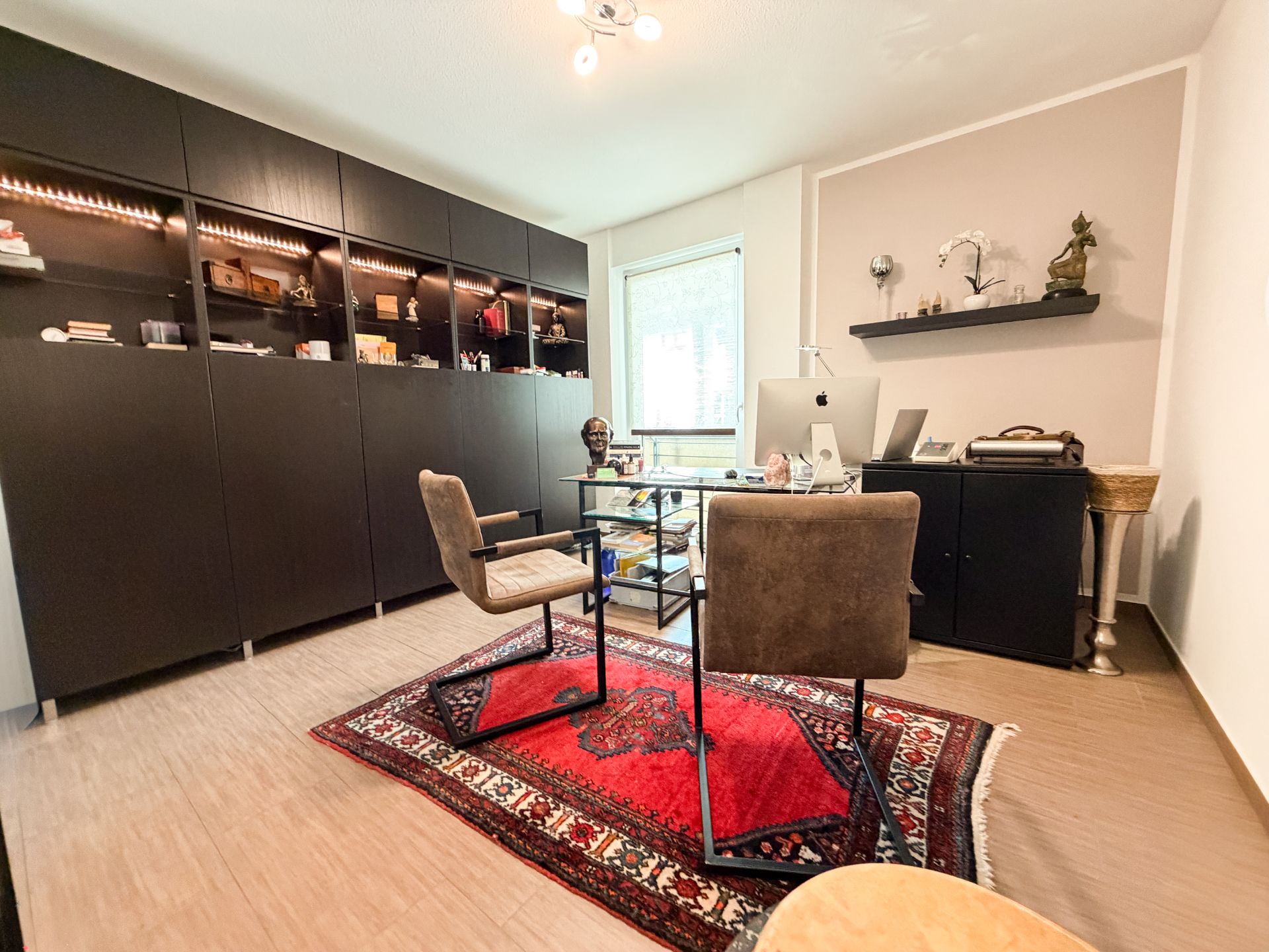 Modernes Arbeitszimmer mit dunklen Regalen, Glas-Schreibtisch und rotem Teppich.
